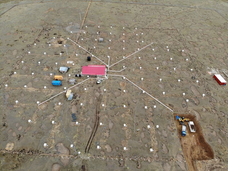 Vista aérea de la meseta de Chacaltaya con decenas de detectores blancos alineados en retícula, canalizaciones radiales, una nave roja en el centro y una excavación con maquinaria en la esquina inferior derecha.