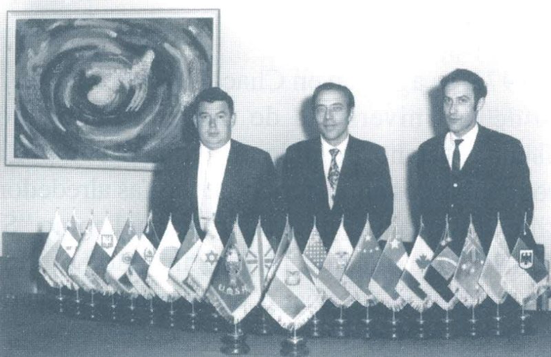 Tres hombres de traje detrás de una fila de banderas de mesa.