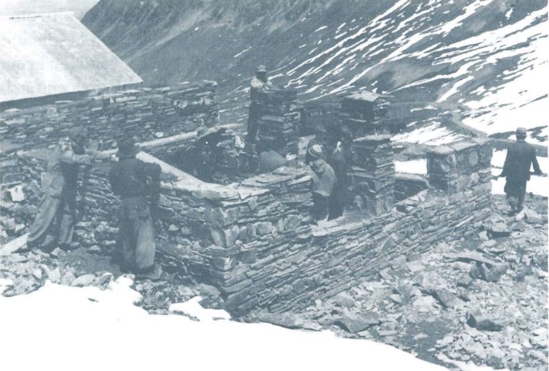 Cinco hombres levantan a mano un muro de piedra en una ladera nevada de alta montaña.