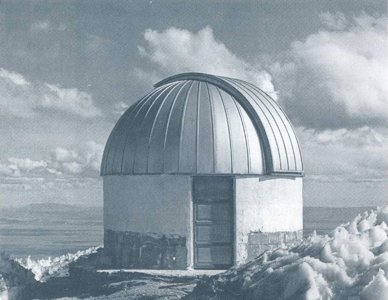 Cúpula metálica de observatorio sobre una base blanca, entre rocas y nieve.