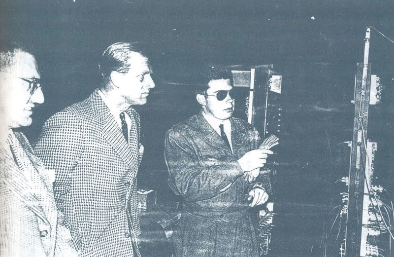 Tres hombres de traje observan un bastidor de instrumentos electrónicos.