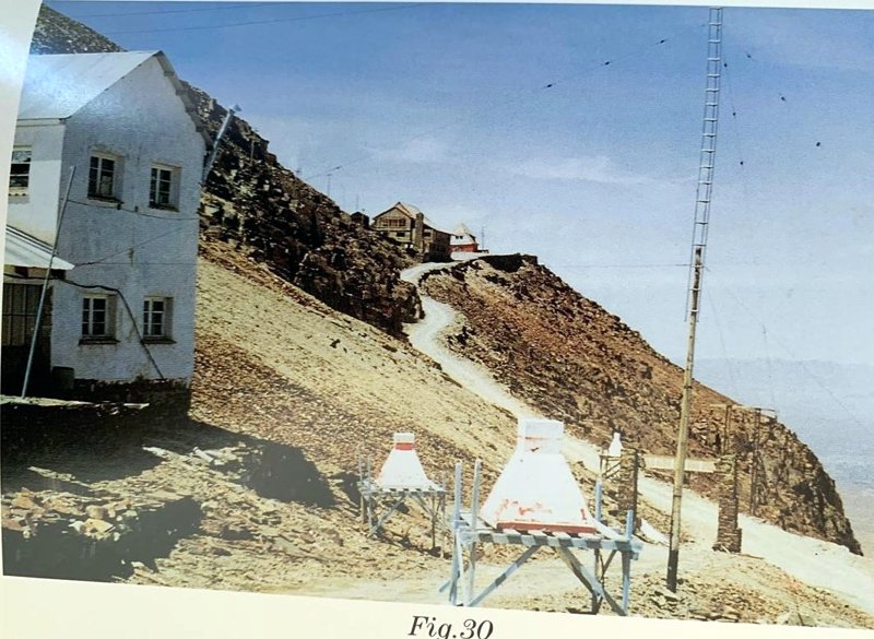 Ladera de la montaña con dos detectores de guía de luz piramidal blanca sobre tripodes, el edificio del Laboratorio a la izquierda y una torre al fondo.