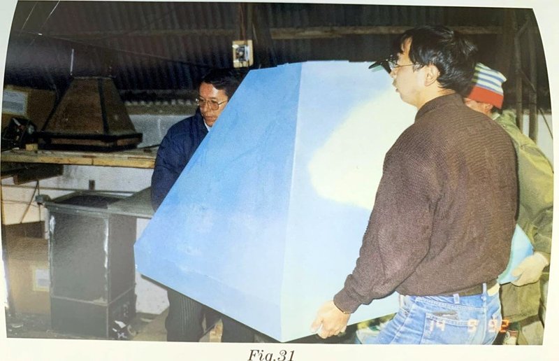 Dos hombres cargan una gran caja piramidal azul dentro de un galpón; sello de fecha del 14 de mayo de 1992.